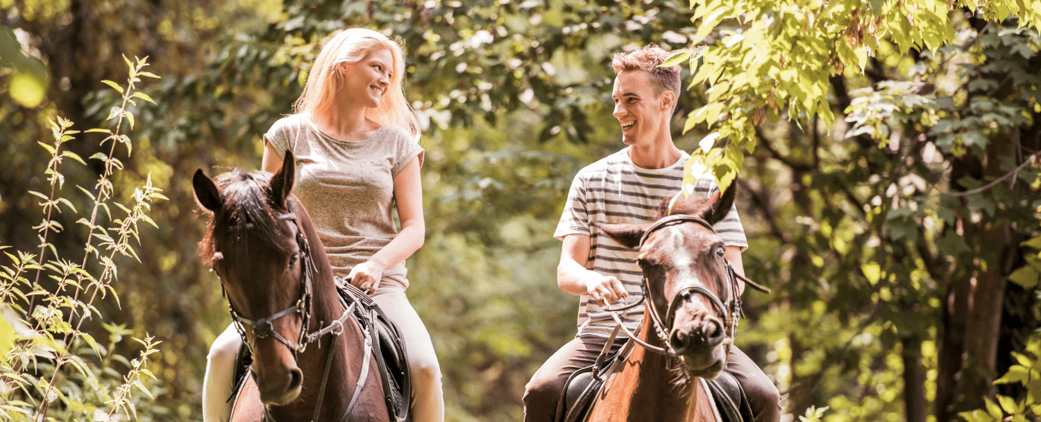 man and woman horseback riding in a wooded area on a sunny day 