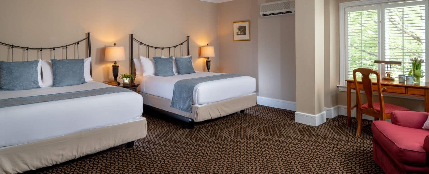 two cozy neat beds next to each other in a large suite with an armchair on the other side and a wooden desk and chair in front of a window