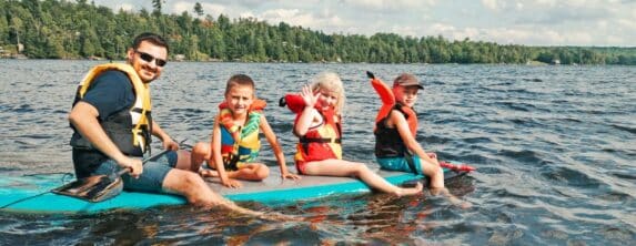 dad and three small children all sitting on a paddle board in the middle of a lake with green tree lined shore behind them