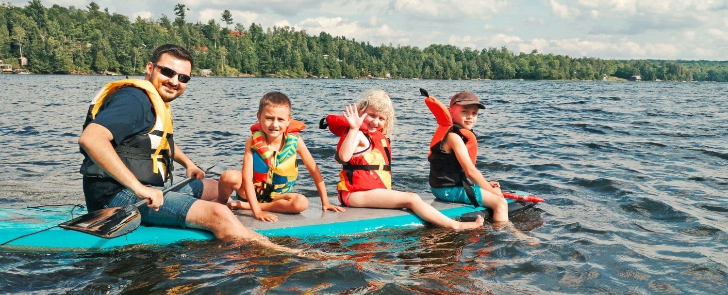 dad and three small children all sitting on a paddle board in the middle of a lake with green tree lined shore behind them