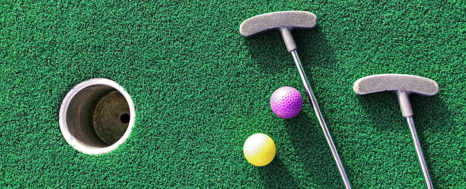 green turf of a mini golf place with a couple of clubs and colored ball next to the pin hole