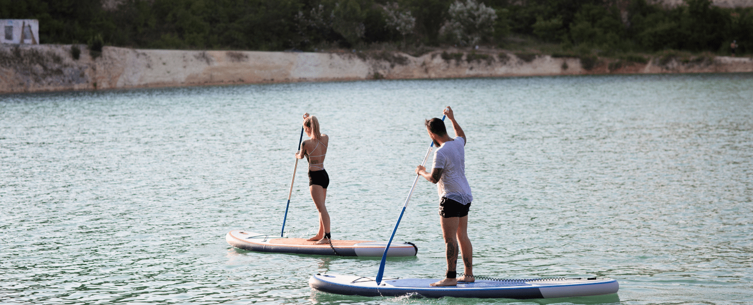 two people standing on paddleboards gliding across a lake water with a rocky shoreline in the background 
