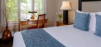 View across bed with blue throw pillows, nightstand with lamp, oak desk with lamp by windown with mini blinds