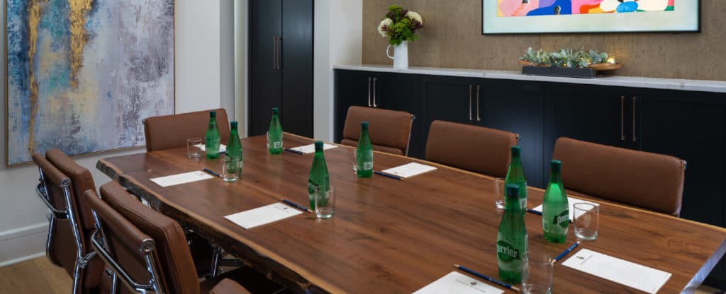 Meeting room with long wooden table holding Perrier waters and notebooks with brown cushioned chairs and white marble topped buffet counter