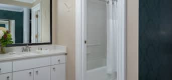 White vanity with flowers in vase, framed miror, white tiled shower and tub combo with grab bar